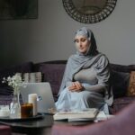 Middle Eastern woman sitting on a sofa working on a laptop in a living room setting.